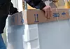 Person hebt ein großes, verpacktes Haushaltsgerät an einer Kartonkante.