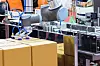 Roboterarm schiebt Kartons auf ein Förderband in einer automatisierten Anlage.
