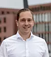 Lächelnder Mann in weißem Hemd vor unscharfen Gebäuden im Hintergrund.